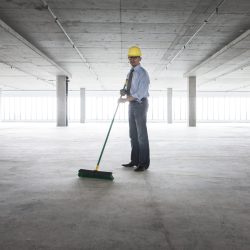 Asian businessman cleaning up with a broom in a large empty raw office space.