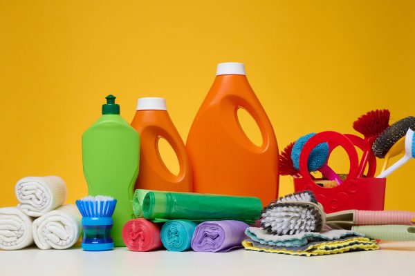 Orange plastic bottles with liquid products and garbage bags, brushes and textile rags for washing floors on a white table, yellow background