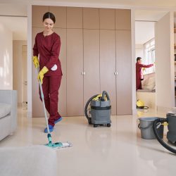 Smiling female janitor washing floor with microfiber dust mop while her colleague cleaning window